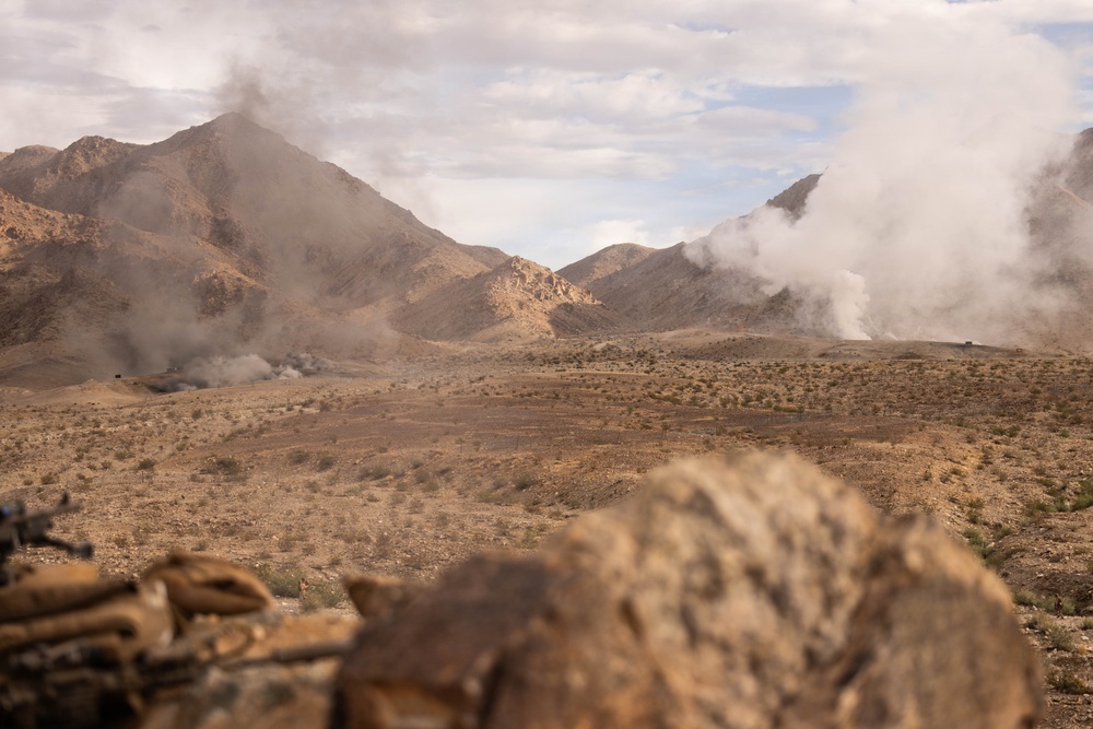 A desert mountain landscape with multiple columns of smoke rising from at least two distinct locations across the terrain. The foreground contains out-of-focus rocks and what appears to be sandbags or equipment, suggesting an observation or defensive position. The scene depicts an arid, sparsely vegetated environment with rugged mountain ridgelines in the background.