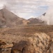 A desert mountain landscape with multiple columns of smoke rising from at least two distinct locations across the terrain. The foreground contains out-of-focus rocks and what appears to be sandbags or equipment, suggesting an observation or defensive position. The scene depicts an arid, sparsely vegetated environment with rugged mountain ridgelines in the background.