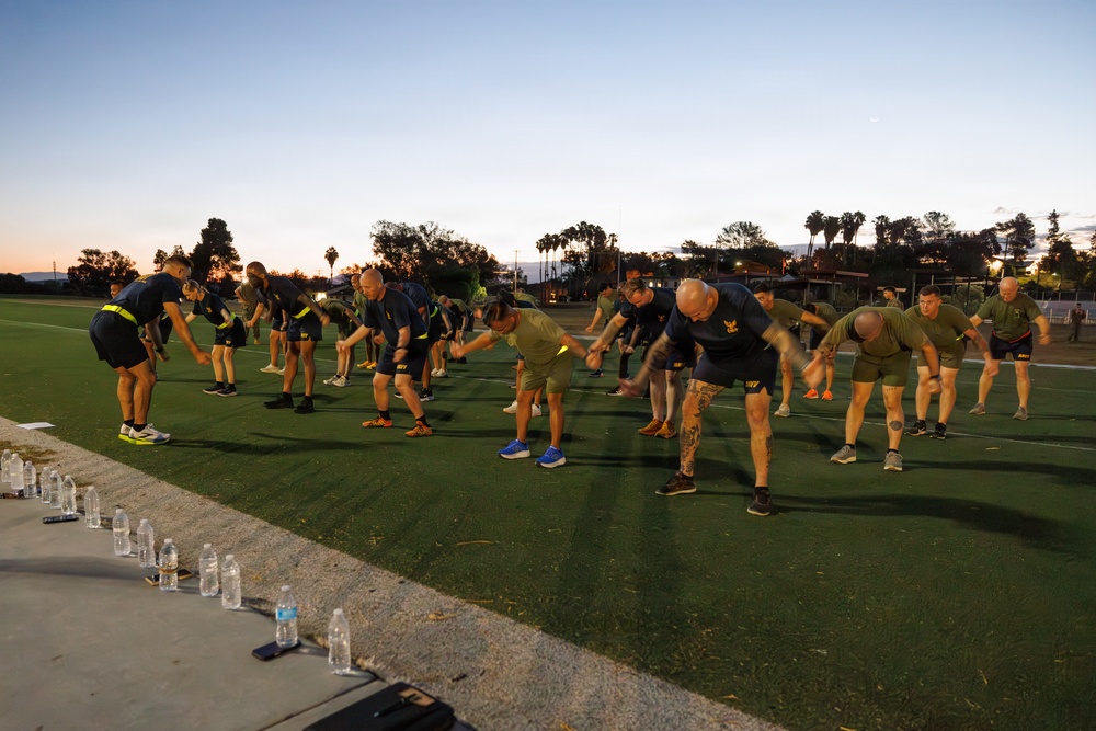 A large group of individuals in matching dark athletic uniforms with yellow accents are performing synchronized stretching or warm-up exercises on a green turf field at dusk or dawn. The group appears to be military or law enforcement personnel based on their uniform consistency and disciplined formation. Water bottles are lined up along the edge of the field, suggesting an organized, supervised physical training session.