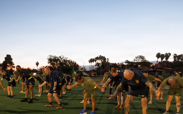 A large group of individuals in matching dark athletic uniforms with yellow accents are performing synchronized stretching or warm-up exercises on a green turf field at dusk or dawn. The group appears to be military or law enforcement personnel based on their uniform consistency and disciplined formation. Water bottles are lined up along the edge of the field, suggesting an organized, supervised physical training session.