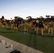 A large group of individuals in matching dark athletic uniforms with yellow accents are performing synchronized stretching or warm-up exercises on a green turf field at dusk or dawn. The group appears to be military or law enforcement personnel based on their uniform consistency and disciplined formation. Water bottles are lined up along the edge of the field, suggesting an organized, supervised physical training session.