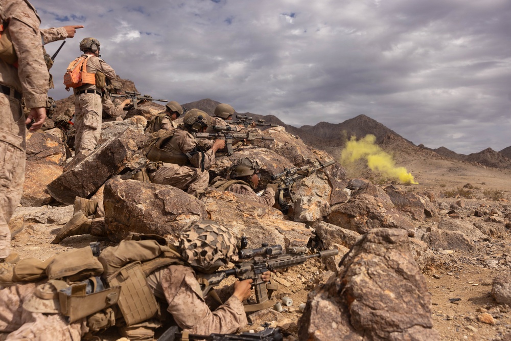 Military personnel in desert camouflage uniforms are positioned behind a rocky outcropping in an arid, mountainous terrain, aiming rifles in a tactical formation. A yellow smoke grenade is visible in the background, suggesting a training exercise or simulated combat scenario. One individual wearing an orange pack appears to be in a supervisory or observer role.
