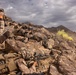 Military personnel in desert camouflage uniforms are positioned behind a rocky outcropping in an arid, mountainous terrain, aiming rifles in a tactical formation. A yellow smoke grenade is visible in the background, suggesting a training exercise or simulated combat scenario. One individual wearing an orange pack appears to be in a supervisory or observer role.