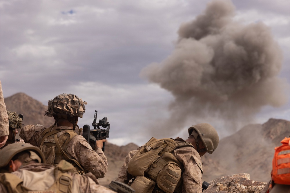 Military personnel in desert camouflage uniforms are taking cover behind rocky terrain while a large smoke plume rises in the background, consistent with an explosion or controlled detonation. At least three soldiers are visible, one of whom is raising a rifle. The arid, mountainous landscape and the presence of an orange safety vest suggest this may be a training exercise.