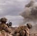 Military personnel in desert camouflage uniforms are taking cover behind rocky terrain while a large smoke plume rises in the background, consistent with an explosion or controlled detonation. At least three soldiers are visible, one of whom is raising a rifle. The arid, mountainous landscape and the presence of an orange safety vest suggest this may be a training exercise.