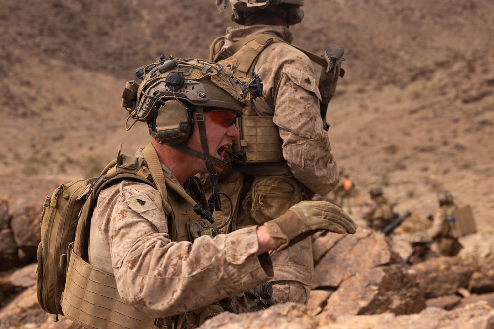 The image shows military personnel in desert camouflage uniforms operating in a rocky, arid terrain. The foreground soldier appears to be shouting and gesturing, likely issuing commands or coordinating with other personnel. Additional soldiers are visible in the background in tactical positions.