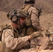 The image shows military personnel in desert camouflage uniforms operating in a rocky, arid terrain. The foreground soldier appears to be shouting and gesturing, likely issuing commands or coordinating with other personnel. Additional soldiers are visible in the background in tactical positions.