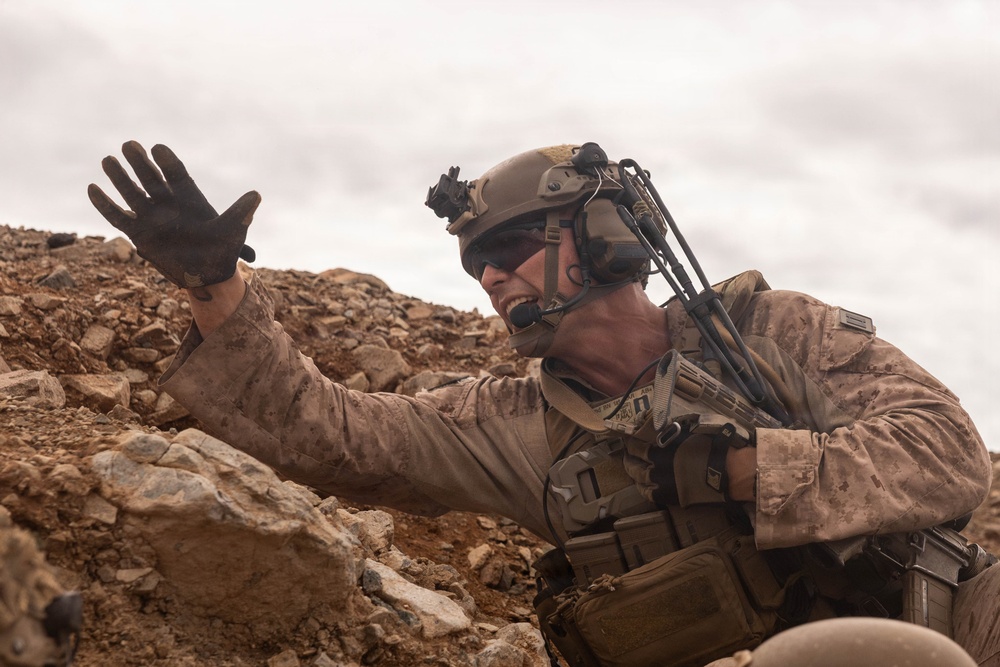 A heavily equipped military service member is positioned low among rocky terrain, raising one gloved hand in what appears to be a hand signal or halt gesture. The soldier is wearing desert camouflage (MARPAT pattern consistent with USMC), a tactical helmet with night-vision mount, communications headset with boom microphone, and a plate carrier with multiple pouches. The rocky, arid landscape and overcast sky suggest a desert operational environment.