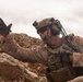 A heavily equipped military service member is positioned low among rocky terrain, raising one gloved hand in what appears to be a hand signal or halt gesture. The soldier is wearing desert camouflage (MARPAT pattern consistent with USMC), a tactical helmet with night-vision mount, communications headset with boom microphone, and a plate carrier with multiple pouches. The rocky, arid landscape and overcast sky suggest a desert operational environment.