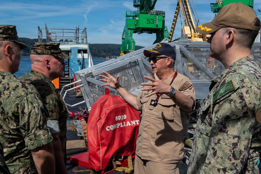 Military personnel in various camouflage uniforms are gathered on what appears to be a naval dock or vessel facility. A central figure wearing tan/khaki Navy uniform and a dark cap with sunglasses is gesturing with both hands, apparently briefing or explaining something to the group. A large green crane numbered '53' is visible in the background along with waterfront infrastructure.
