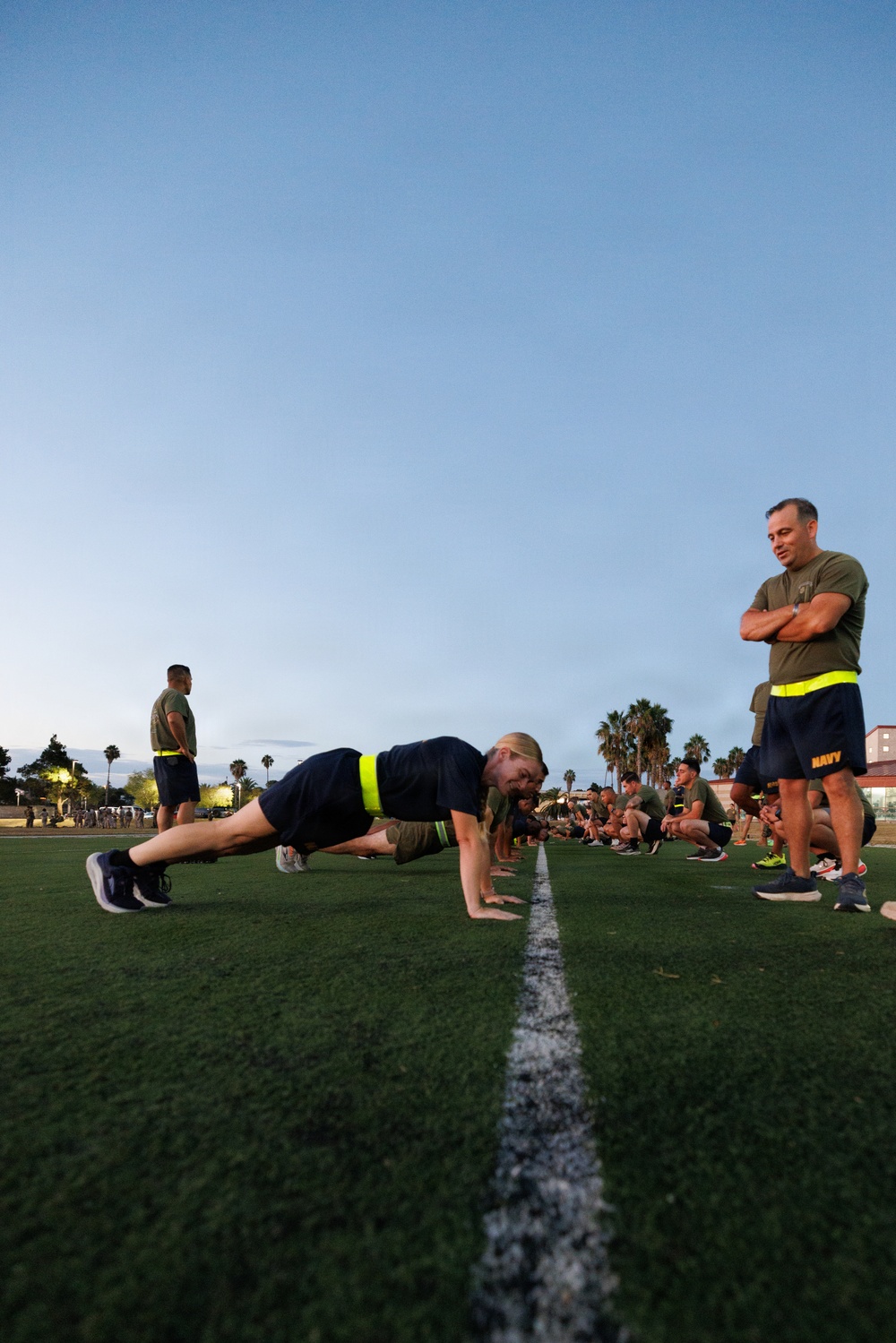 A military physical training session taking place on a turf athletic field at dusk or early morning. Personnel in Navy/military PT gear are performing push-ups along a field line while an observer with arms crossed watches. Multiple service members are visible in the background in various exercise positions.
