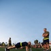 A military physical training session taking place on a turf athletic field at dusk or early morning. Personnel in Navy/military PT gear are performing push-ups along a field line while an observer with arms crossed watches. Multiple service members are visible in the background in various exercise positions.