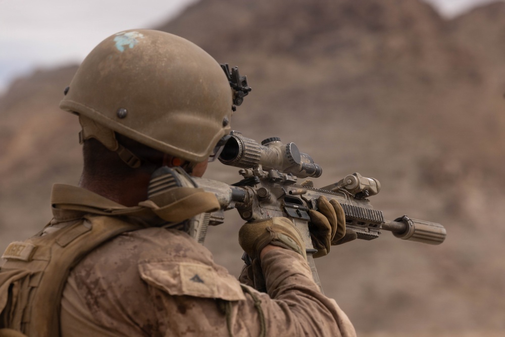 A soldier in desert camouflage uniform and tactical helmet is aiming a rifle equipped with an optical scope in an arid, mountainous environment. The image is taken from behind and slightly to the side, showing the soldier in a shooting stance. The setting and equipment suggest a military training exercise or operational deployment in a desert region.