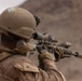A soldier in desert camouflage uniform and tactical helmet is aiming a rifle equipped with an optical scope in an arid, mountainous environment. The image is taken from behind and slightly to the side, showing the soldier in a shooting stance. The setting and equipment suggest a military training exercise or operational deployment in a desert region.