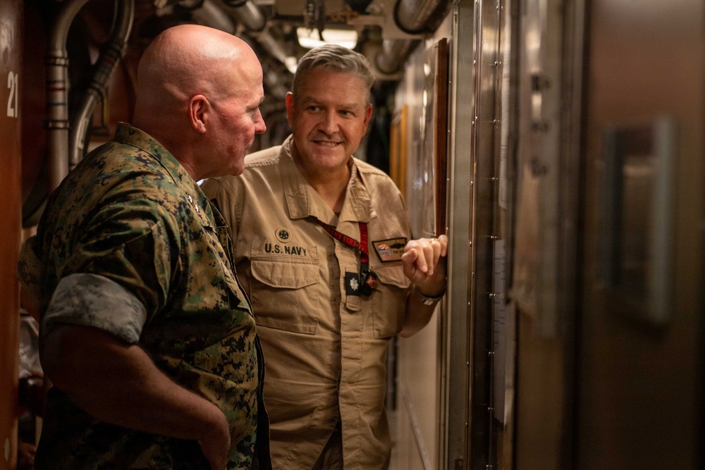 Two military personnel are standing in a narrow corridor, likely aboard a naval vessel or military facility. One individual is wearing U.S. Marine Corps digital camouflage (MARPAT) uniform, while the other is wearing a tan U.S. Navy coverall uniform. They appear to be reviewing or discussing something on a display board mounted on the wall.