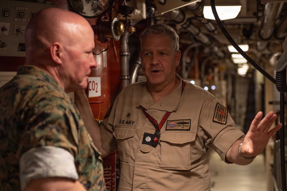 Two military personnel are engaged in conversation inside what appears to be a naval vessel, likely a submarine or ship interior. One individual is wearing U.S. Marine Corps camouflage (MARPAT) uniform, while the other is in U.S. Navy khaki coveralls with a name tag reading 'Ryan Grundt' and the designation 'CO' (Commanding Officer). The Navy officer appears to be briefing or explaining something, gesturing with his right hand.