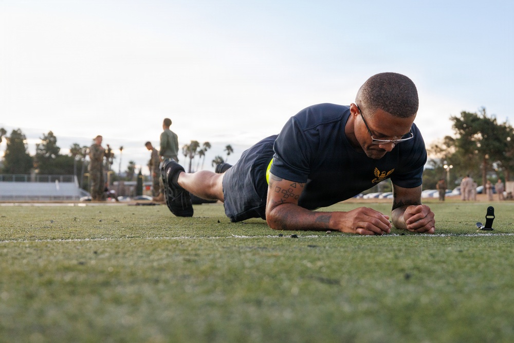 A military service member in Navy physical training attire performs a forearm plank exercise on an artificial turf field. The individual is wearing glasses and has visible tattoos on their arm, with a gold eagle insignia on their shirt indicating Navy rank or rating. In the background, other personnel in military uniforms are visible, suggesting a group physical fitness assessment or training event.