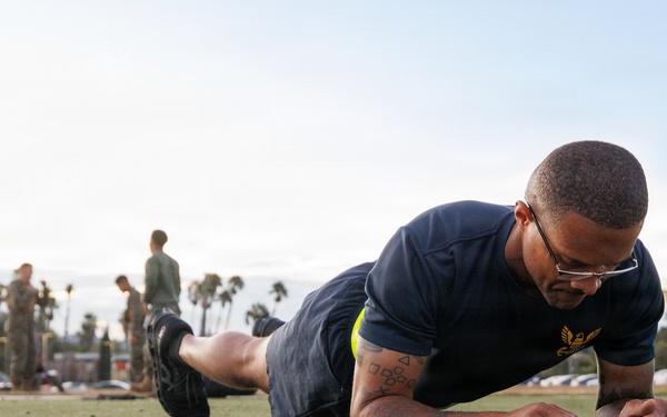 A military service member in Navy physical training attire performs a forearm plank exercise on an artificial turf field. The individual is wearing glasses and has visible tattoos on their arm, with a gold eagle insignia on their shirt indicating Navy rank or rating. In the background, other personnel in military uniforms are visible, suggesting a group physical fitness assessment or training event.