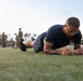 A military service member in Navy physical training attire performs a forearm plank exercise on an artificial turf field. The individual is wearing glasses and has visible tattoos on their arm, with a gold eagle insignia on their shirt indicating Navy rank or rating. In the background, other personnel in military uniforms are visible, suggesting a group physical fitness assessment or training event.