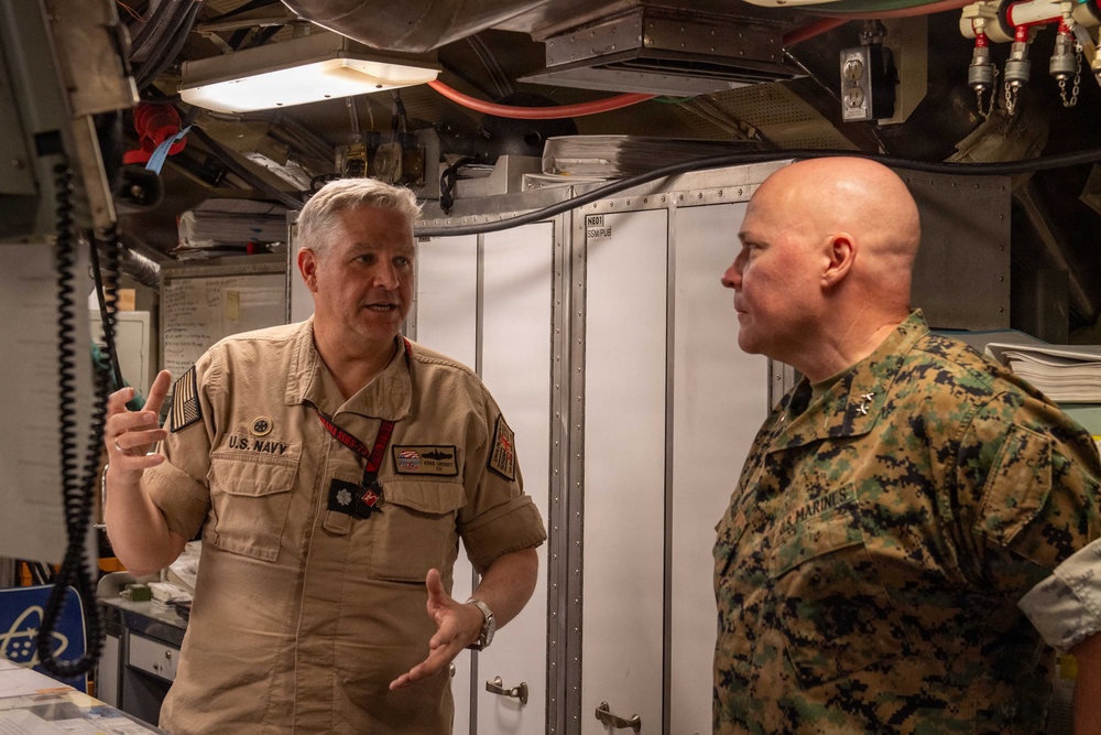 Two U.S. military officers are engaged in conversation inside what appears to be a naval vessel or military facility. The officer on the left is wearing a U.S. Navy tan/khaki uniform and appears to be briefing or explaining something, while the officer on the right is wearing U.S. Marine Corps MARPAT camouflage uniform with general officer rank insignia. The setting appears to be a confined operational space with equipment, cabinets, and overhead utilities visible.