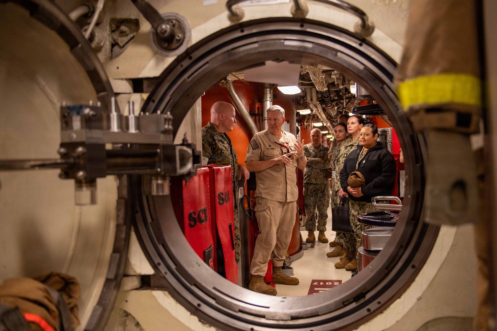 Military personnel are gathered inside what appears to be a naval vessel or submarine, viewed through a large circular watertight hatch/bulkhead door. A senior officer in tan/khaki uniform appears to be briefing or conducting a tour for a group of Navy and Marine Corps personnel. The framing through the circular hatch creates a distinctive compositional perspective of the interior compartment.