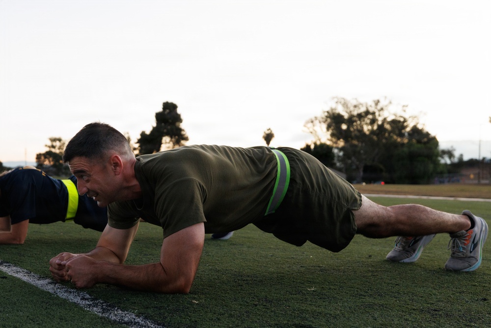 A man in military-style olive green athletic wear is performing a plank exercise on a grass athletic field during early morning or evening hours. A second individual in dark clothing with a yellow-green reflective vest is partially visible to the left, performing the same exercise. The setting appears to be a military or organized fitness training session on a turf field.