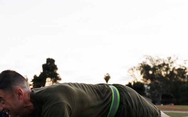 A man in military-style olive green athletic wear is performing a plank exercise on a grass athletic field during early morning or evening hours. A second individual in dark clothing with a yellow-green reflective vest is partially visible to the left, performing the same exercise. The setting appears to be a military or organized fitness training session on a turf field.