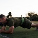 A man in military-style olive green athletic wear is performing a plank exercise on a grass athletic field during early morning or evening hours. A second individual in dark clothing with a yellow-green reflective vest is partially visible to the left, performing the same exercise. The setting appears to be a military or organized fitness training session on a turf field.
