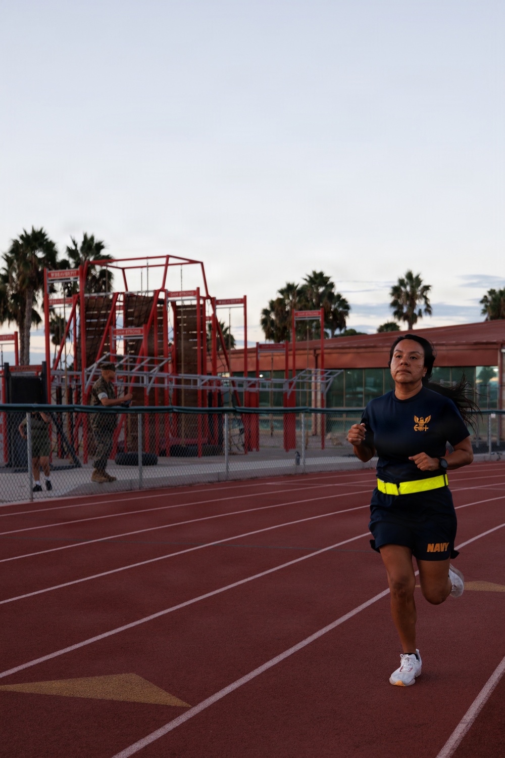 A female U.S. Navy service member is running on a red athletic track, wearing standard Navy PT (physical training) gear including a dark blue shirt with a gold eagle insignia, navy shorts labeled 'NAVY', and a high-visibility yellow belt. In the background, a red military obstacle course structure is visible along with palm trees and another uniformed individual near a chain-link fence. The setting appears to be a military installation, likely in a warm climate, photographed during early morning or evening light.