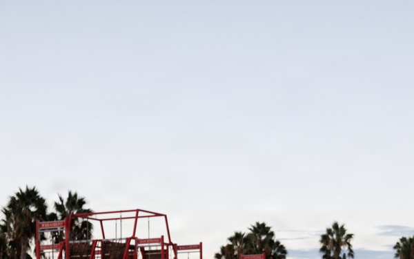 A female U.S. Navy service member is running on a red athletic track, wearing standard Navy PT (physical training) gear including a dark blue shirt with a gold eagle insignia, navy shorts labeled 'NAVY', and a high-visibility yellow belt. In the background, a red military obstacle course structure is visible along with palm trees and another uniformed individual near a chain-link fence. The setting appears to be a military installation, likely in a warm climate, photographed during early morning or evening light.