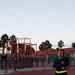 A female U.S. Navy service member is running on a red athletic track, wearing standard Navy PT (physical training) gear including a dark blue shirt with a gold eagle insignia, navy shorts labeled 'NAVY', and a high-visibility yellow belt. In the background, a red military obstacle course structure is visible along with palm trees and another uniformed individual near a chain-link fence. The setting appears to be a military installation, likely in a warm climate, photographed during early morning or evening light.