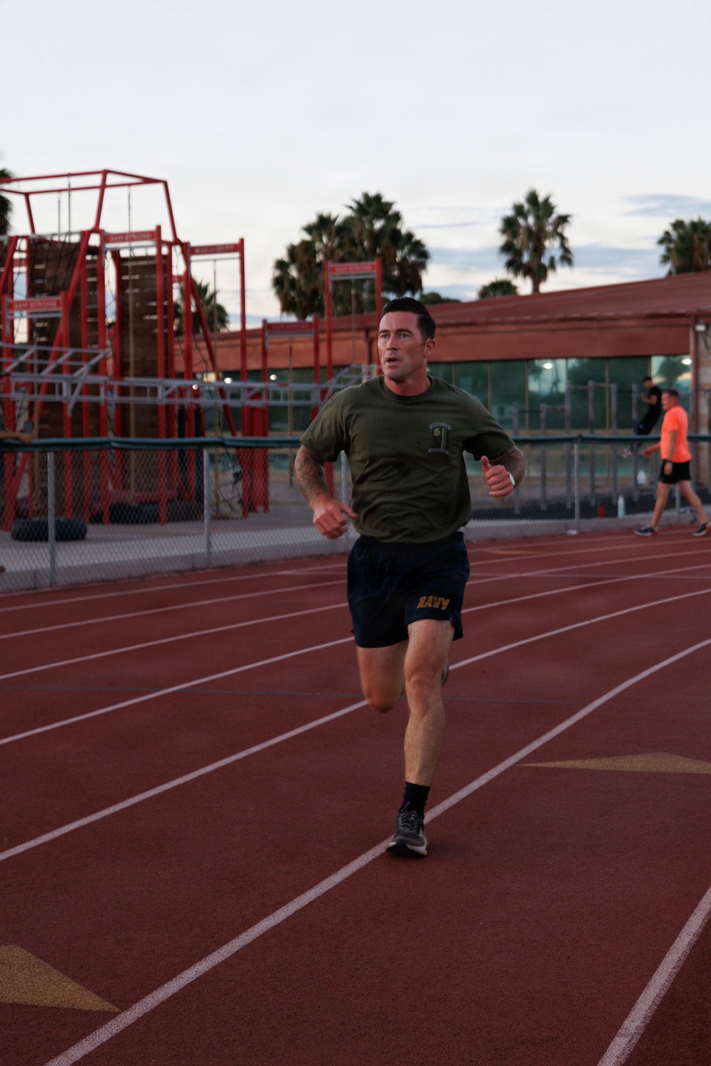 A man wearing a military-style olive green t-shirt and navy blue 'NAVY' shorts is running on a red athletic track. The setting appears to be an outdoor sports facility with bleachers, chain-link fencing, and palm trees visible in the background. The photo was taken during early morning or evening hours based on the soft, low-light sky conditions.