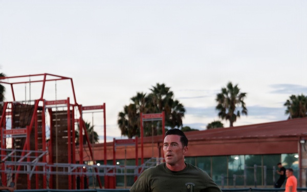 A man wearing a military-style olive green t-shirt and navy blue 'NAVY' shorts is running on a red athletic track. The setting appears to be an outdoor sports facility with bleachers, chain-link fencing, and palm trees visible in the background. The photo was taken during early morning or evening hours based on the soft, low-light sky conditions.