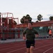 A man wearing a military-style olive green t-shirt and navy blue 'NAVY' shorts is running on a red athletic track. The setting appears to be an outdoor sports facility with bleachers, chain-link fencing, and palm trees visible in the background. The photo was taken during early morning or evening hours based on the soft, low-light sky conditions.