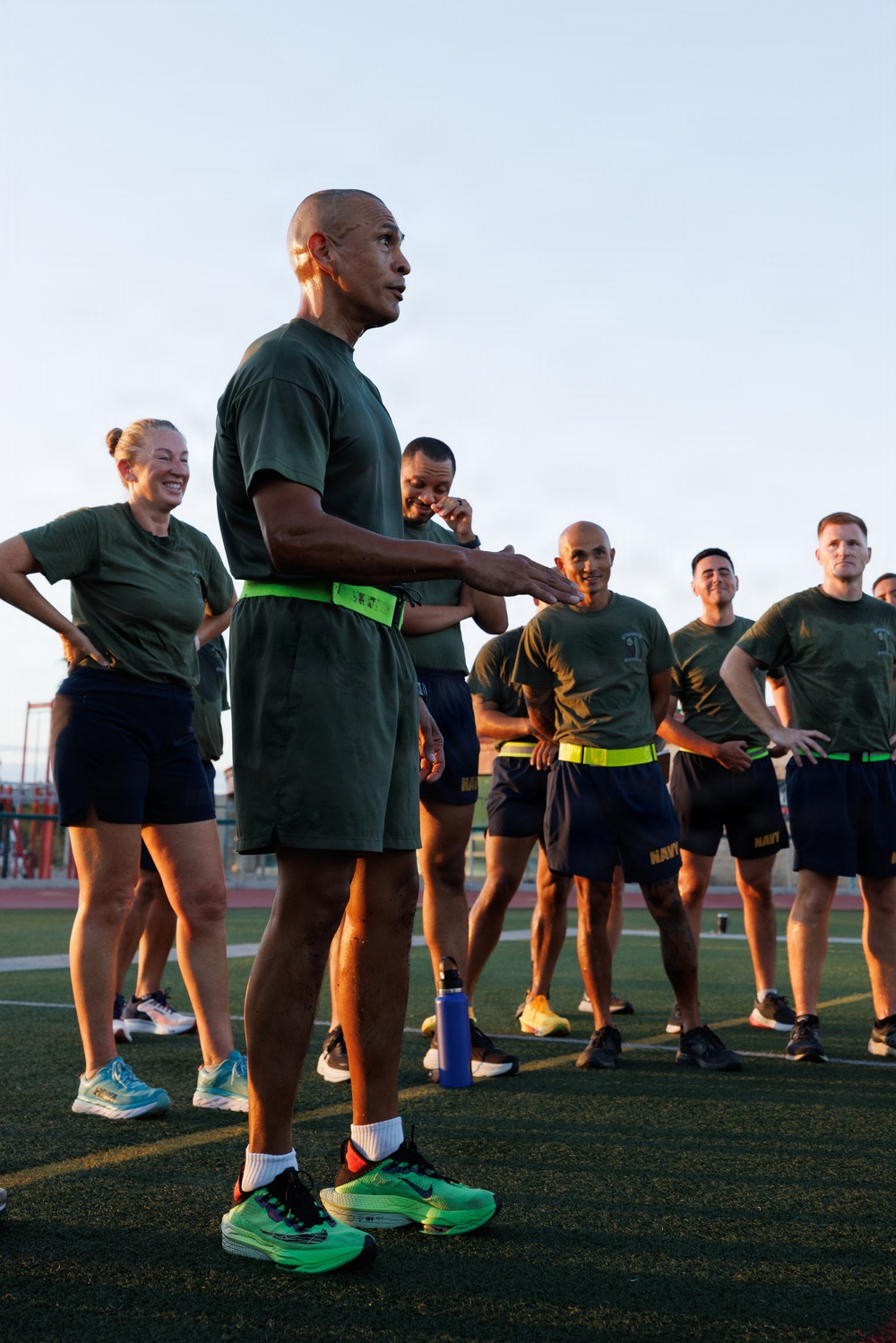A group of military personnel in physical training (PT) attire are gathered on an athletic field or track. A central figure, wearing a green t-shirt, dark shorts, and bright green Nike running shoes with a neon green waist pack, appears to be addressing or briefing the group. Several members are wearing shorts with 'NAVY' printed on them, suggesting this is a U.S. Navy PT session.