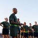 A group of military personnel in physical training (PT) attire are gathered on an athletic field or track. A central figure, wearing a green t-shirt, dark shorts, and bright green Nike running shoes with a neon green waist pack, appears to be addressing or briefing the group. Several members are wearing shorts with 'NAVY' printed on them, suggesting this is a U.S. Navy PT session.