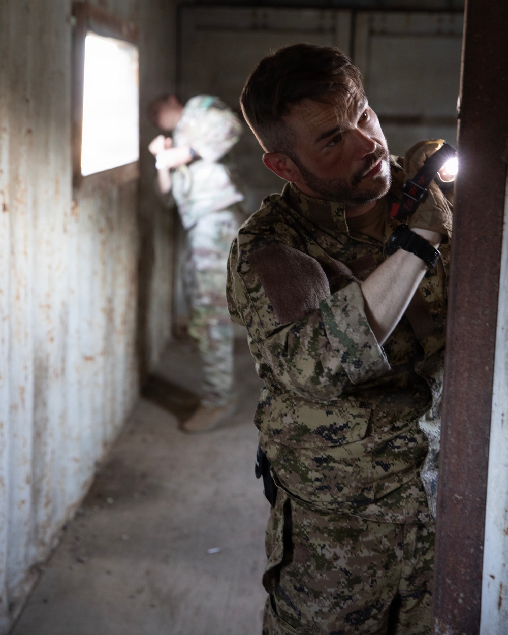 Two military personnel in camouflage uniforms are conducting a building clearance operation inside a deteriorated structure. The foreground soldier is using a tactical flashlight held in a high-ready position while pressing against a wall near a doorway. A second soldier is visible in the background near a window, appearing to maintain a security position.