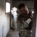 Two military personnel in camouflage uniforms are conducting a building clearance operation inside a deteriorated structure. The foreground soldier is using a tactical flashlight held in a high-ready position while pressing against a wall near a doorway. A second soldier is visible in the background near a window, appearing to maintain a security position.