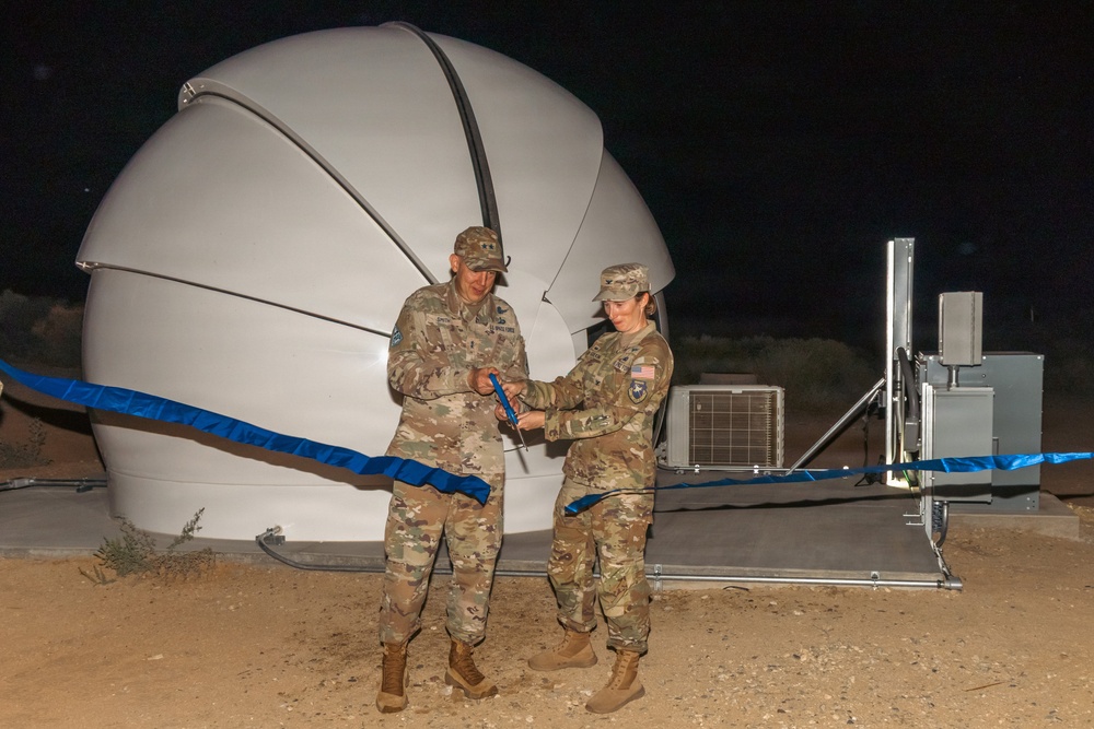 Two military personnel in camouflage uniforms are performing a ribbon-cutting ceremony at night in front of a large white radome (dome-shaped antenna enclosure). The ceremony appears to mark the inauguration or activation of a satellite communications or radar installation. Both individuals are holding scissors and cutting a blue ribbon stretched across the facility.