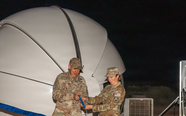 Two military personnel in camouflage uniforms are performing a ribbon-cutting ceremony at night in front of a large white radome (dome-shaped antenna enclosure). The ceremony appears to mark the inauguration or activation of a satellite communications or radar installation. Both individuals are holding scissors and cutting a blue ribbon stretched across the facility.