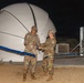 Two military personnel in camouflage uniforms are performing a ribbon-cutting ceremony at night in front of a large white radome (dome-shaped antenna enclosure). The ceremony appears to mark the inauguration or activation of a satellite communications or radar installation. Both individuals are holding scissors and cutting a blue ribbon stretched across the facility.