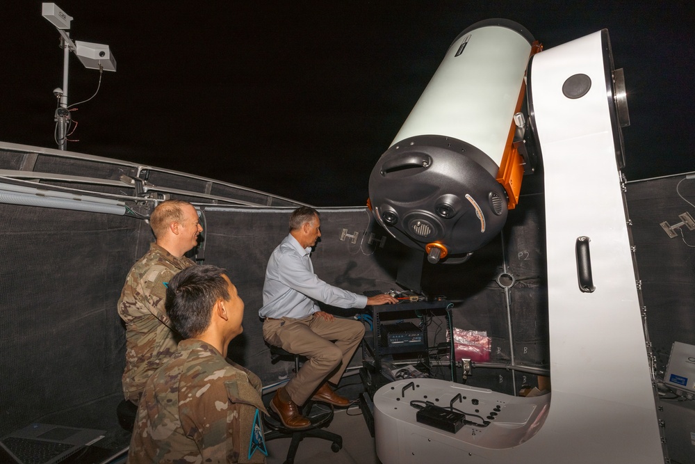 Three individuals are gathered around a large telescope system inside what appears to be an observatory dome or enclosure at night. One civilian in a light blue shirt is seated at a control workstation operating the equipment, while two military personnel in camouflage uniforms observe. The telescope is a large, professional-grade instrument mounted on an orange and white motorized mount.