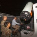 Three individuals are gathered around a large telescope system inside what appears to be an observatory dome or enclosure at night. One civilian in a light blue shirt is seated at a control workstation operating the equipment, while two military personnel in camouflage uniforms observe. The telescope is a large, professional-grade instrument mounted on an orange and white motorized mount.