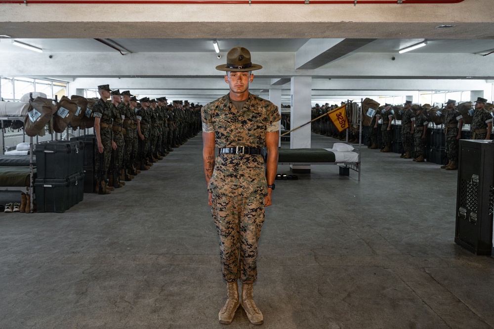 A U.S. Marine drill instructor stands at attention in the center of a military barracks bay, wearing MARPAT digital camouflage utilities and the distinctive campaign cover (drill instructor hat). Rows of recruits in similar uniforms stand at attention along both sides of the bay, with bunk beds, footlockers, and military gear visible in the background. A yellow guidon flag is visible on the right side of the formation.
