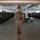Lima Company Senior Drill Instructor Inspection A U.S. Marine drill instructor stands at attention in the center of a military barracks bay, wearing MARPAT digital camouflage utilities and the distinctive campaign cover (drill instructor hat). Rows of recruits in similar uniforms stand at attention along both sides of the bay, with bunk beds, footlockers, and military gear visible in the background. A yellow guidon flag is visible on the right side of the formation.