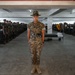 A U.S. Marine drill instructor stands at attention in the center of a military barracks bay, wearing MARPAT digital camouflage utilities and the distinctive campaign cover (drill instructor hat). Rows of recruits in similar uniforms stand at attention along both sides of the bay, with bunk beds, footlockers, and military gear visible in the background. A yellow guidon flag is visible on the right side of the formation.