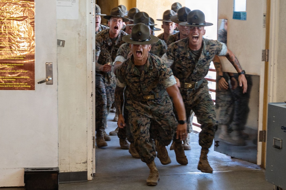 A group of U.S. Marine Corps Drill Instructors in woodland MARPAT camouflage uniforms and campaign covers (Smokey Bear hats) are rushing through a doorway while shouting aggressively. This appears to be a recruit receiving or 'pick-up' event at a Marine Corps Recruit Depot, where drill instructors storm the barracks to intimidate new recruits. The image captures the high-intensity, confrontational nature of Marine Corps basic training initiation.