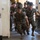 Lima Company Senior Drill Instructor Inspection A group of U.S. Marine Corps Drill Instructors in woodland MARPAT camouflage uniforms and campaign covers (Smokey Bear hats) are rushing through a doorway while shouting aggressively. This appears to be a recruit receiving or 'pick-up' event at a Marine Corps Recruit Depot, where drill instructors storm the barracks to intimidate new recruits. The image captures the high-intensity, confrontational nature of Marine Corps basic training initiation.