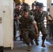 A group of U.S. Marine Corps Drill Instructors in woodland MARPAT camouflage uniforms and campaign covers (Smokey Bear hats) are rushing through a doorway while shouting aggressively. This appears to be a recruit receiving or 'pick-up' event at a Marine Corps Recruit Depot, where drill instructors storm the barracks to intimidate new recruits. The image captures the high-intensity, confrontational nature of Marine Corps basic training initiation.