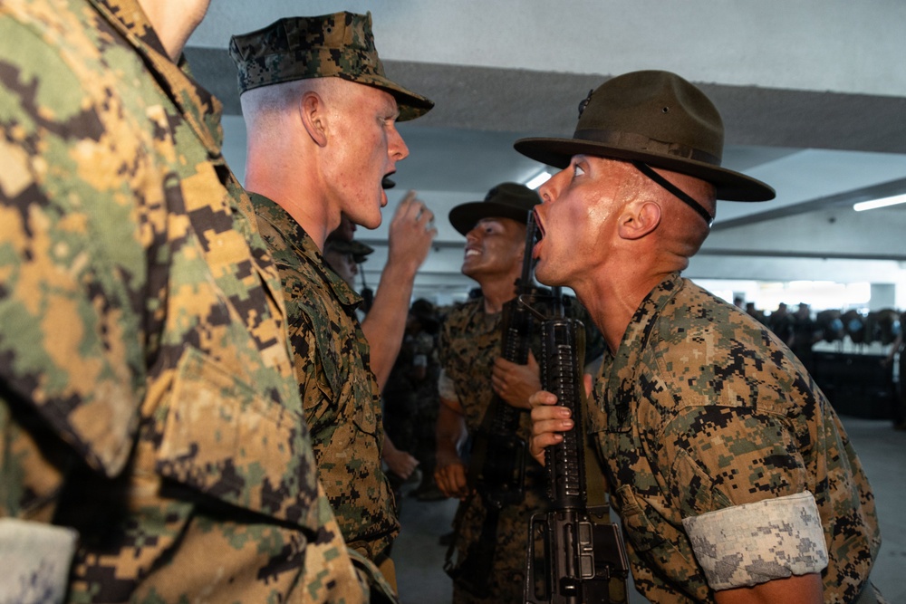 The image depicts a U.S. Marine Corps drill instructor in a campaign cover (campaign hat) shouting face-to-face at a recruit during what appears to be basic training or boot camp. The drill instructor is holding a rifle and wearing a white armband, a common identifier for drill instructors. Multiple other drill instructors and recruits are visible in the background in a covered outdoor or indoor facility.